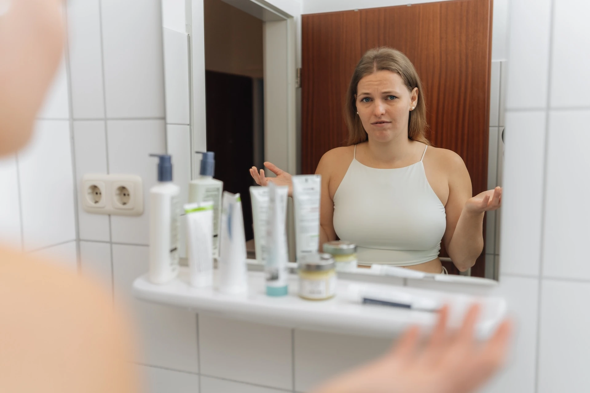 Kristin Eifler von Neurodermitis Selbstheilung im Bad vor dem Spiegel mit Fragendem Blick und einer Auswahl an Cremes und Salben. Sie fragt sich: Welche Creme gegen Neurodermitis?