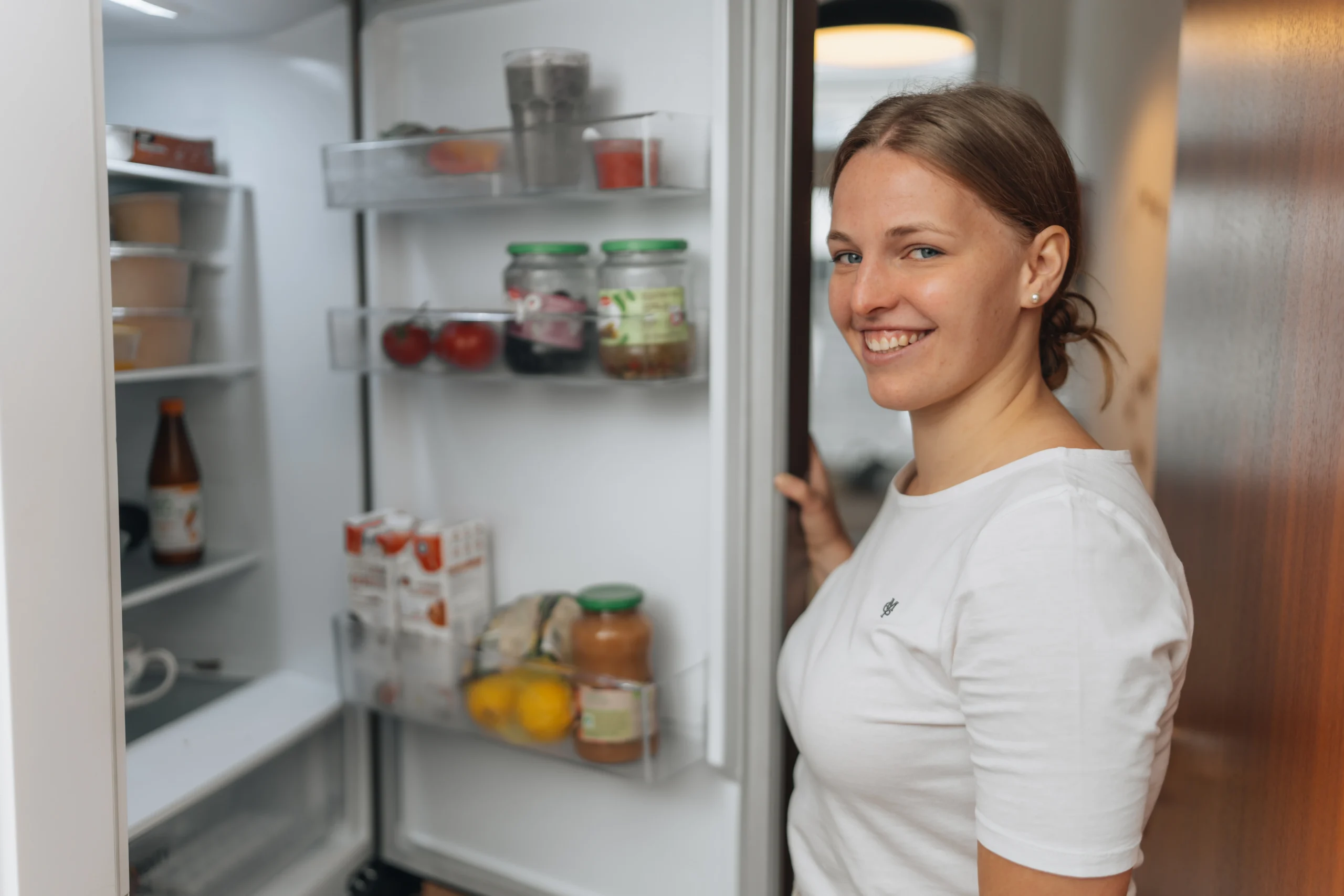 Lächelnde junge Frau öffnet einen Kühlschrank mit Gläsern, Tomaten und Milchtüten in der Tür