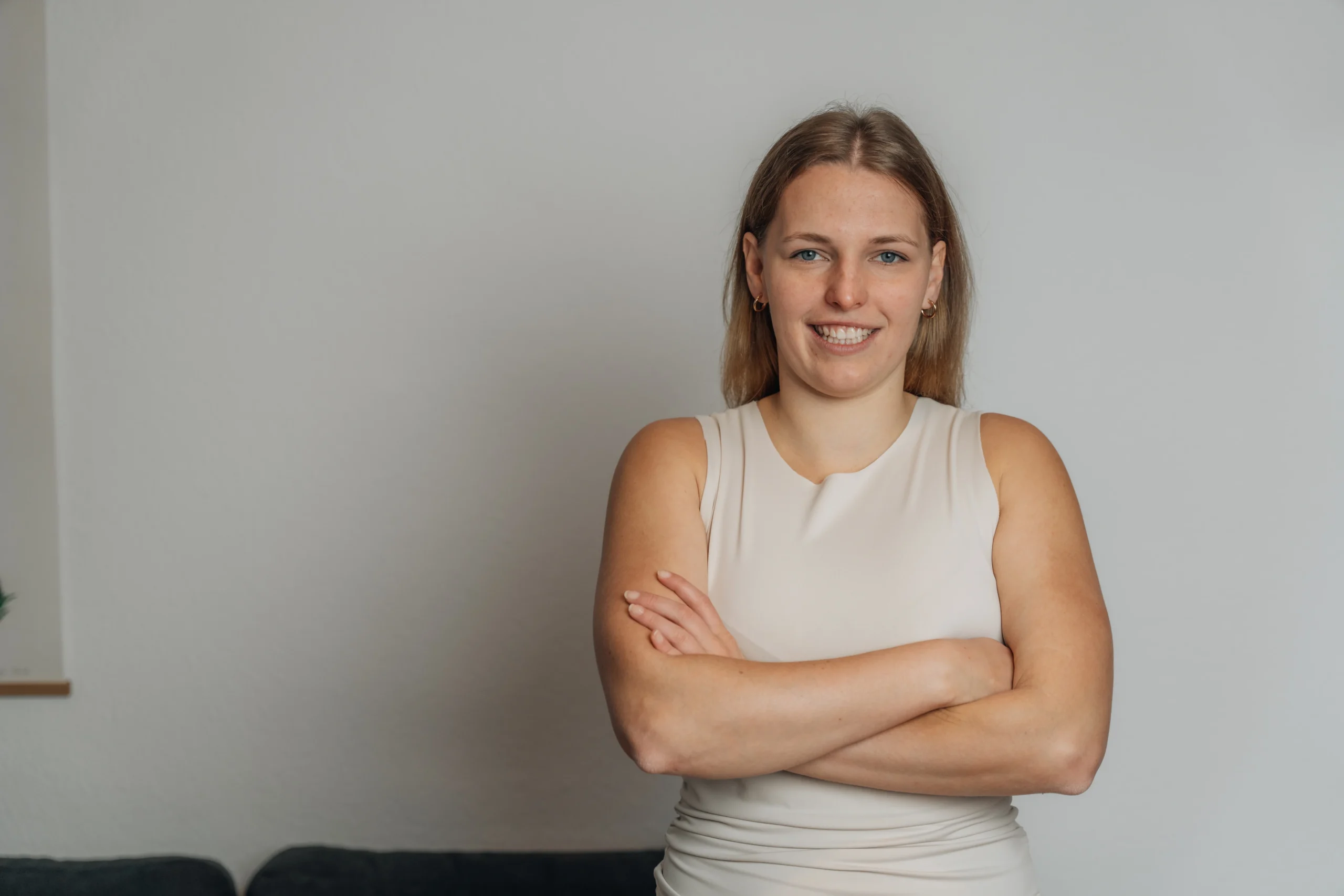 neurodermitis-baby-oberschenkel.webp Schreibe einen kurzen Alt-Text für dieses Bild. Lächelnde junge Frau steht mit verschränkten Armen vor einer hellen Wand