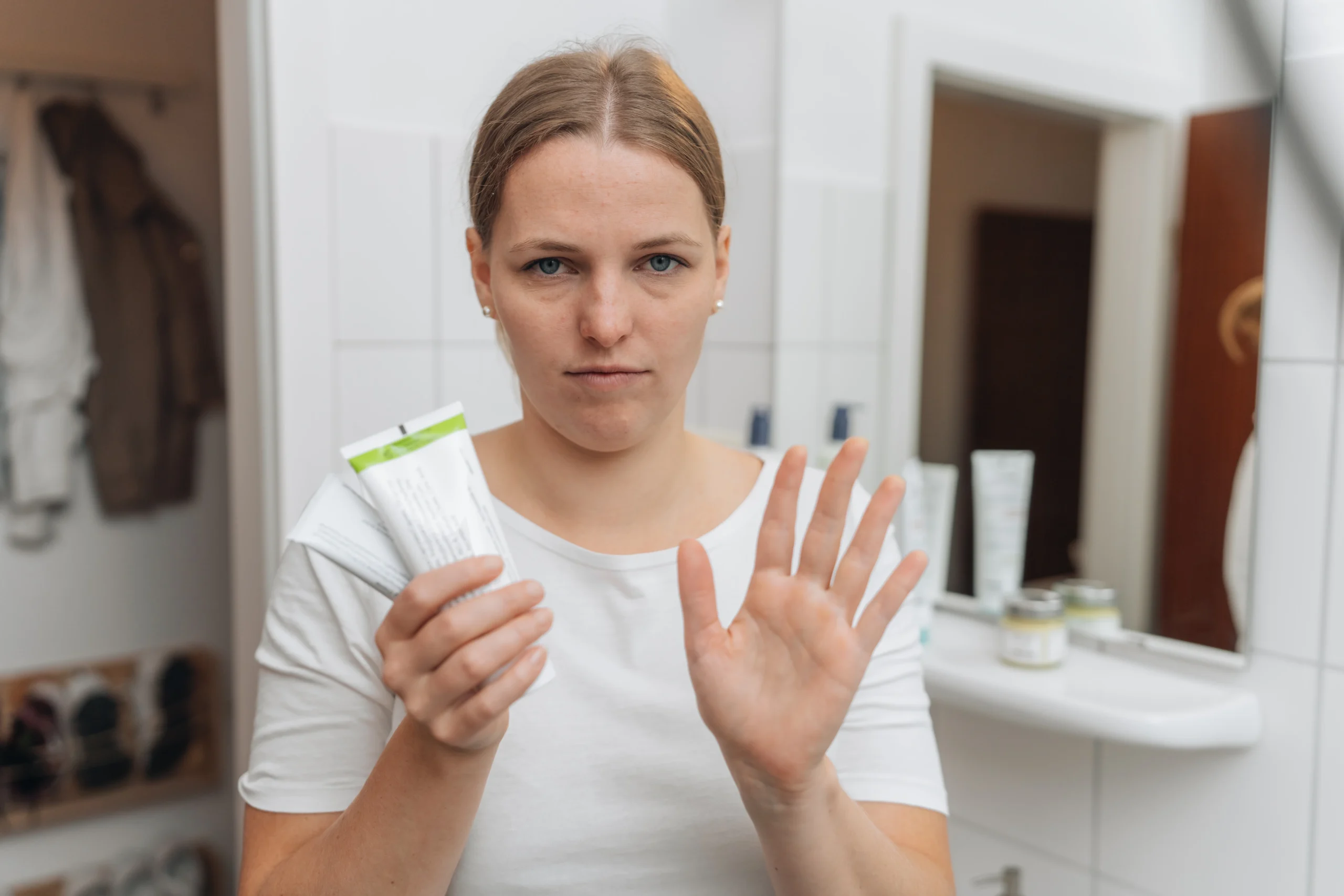 Junge Frau hält im Badezimmer eine Cremetube in der Hand und hebt die andere Hand zu einer Stopp-Geste