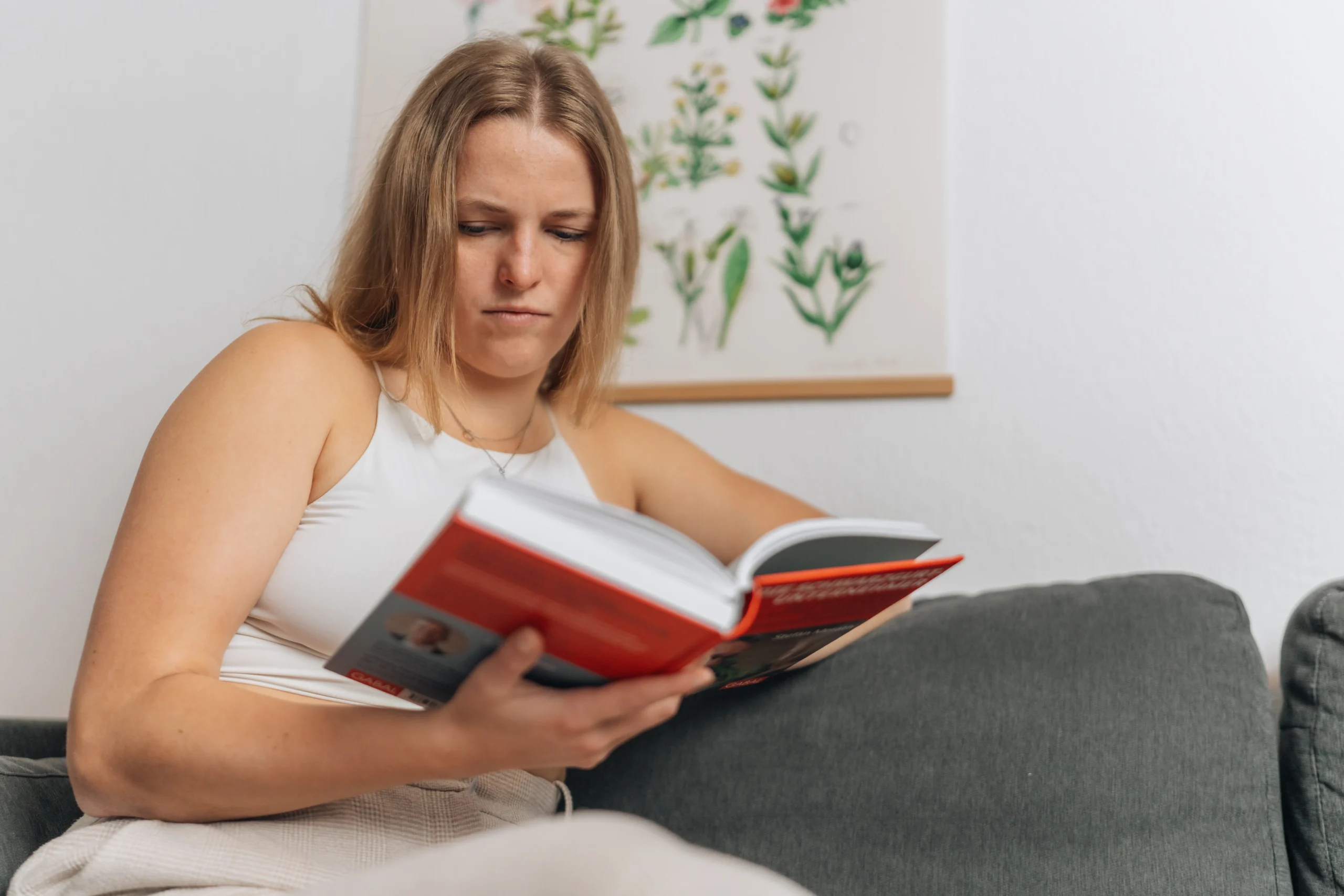 Junge Frau sitzt auf einem Sofa und liest nachdenklich in einem Buch
