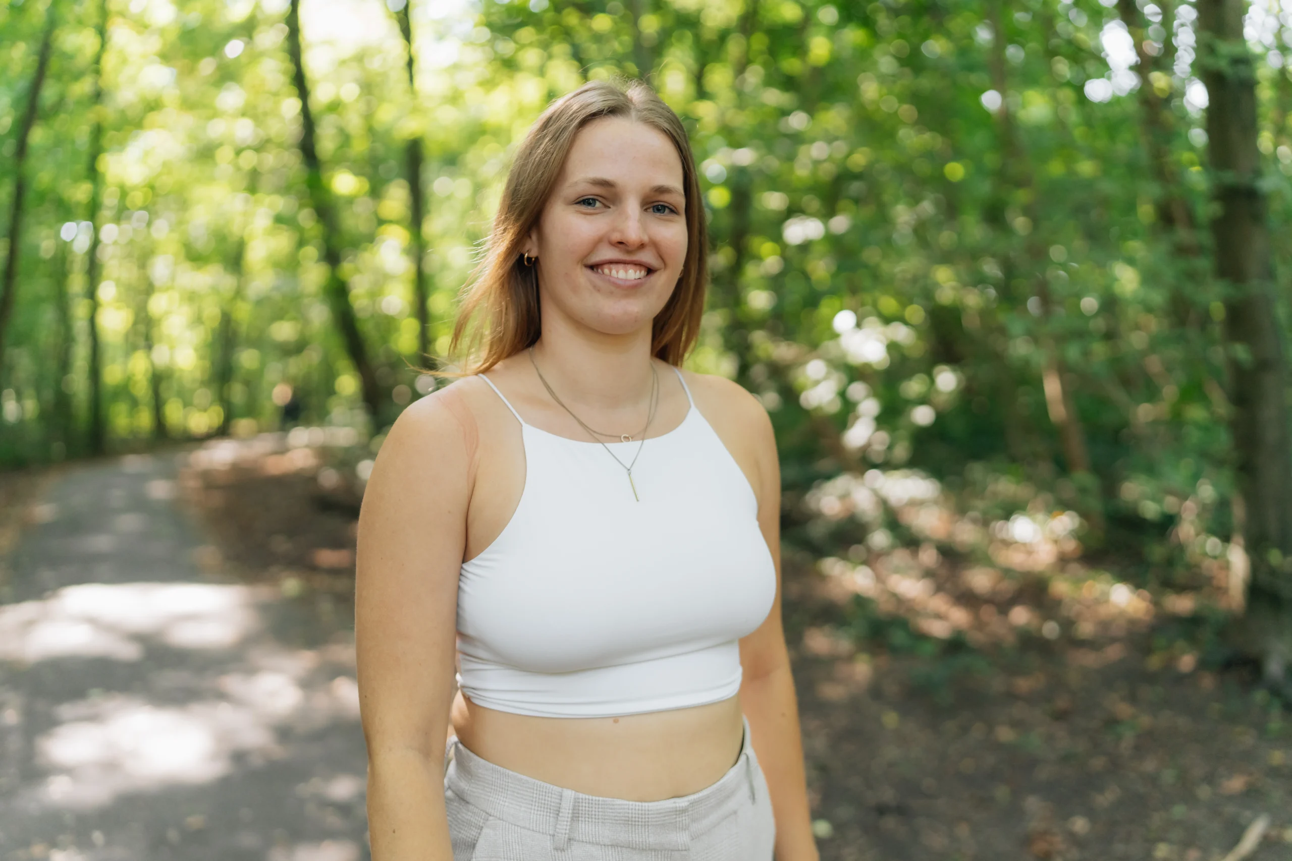 Junge Frau lächelnd auf einem sonnigen Waldweg, in weißem Top und heller Stoffhose